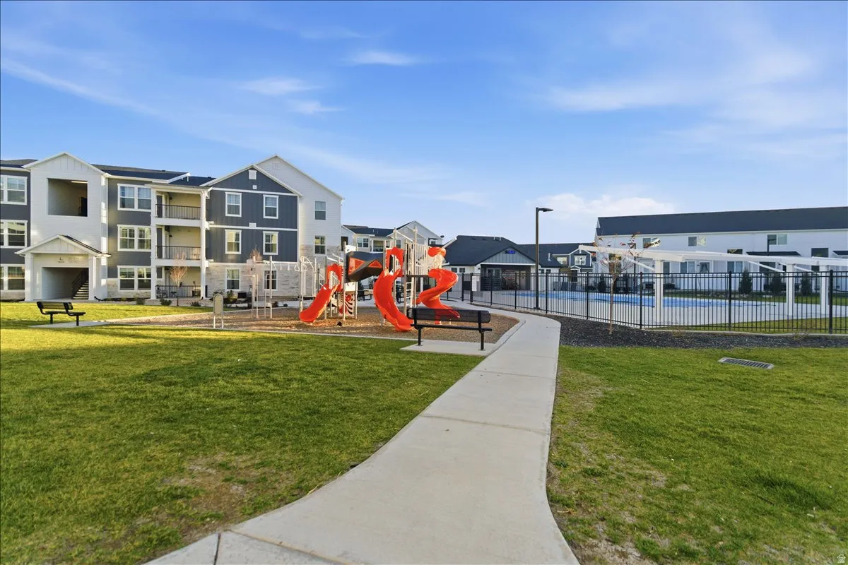 Expansive green space featuring a playground with red slides, a concrete pathway, a fenced swimming pool with a pergola, and multi-story residential buildings with balconies