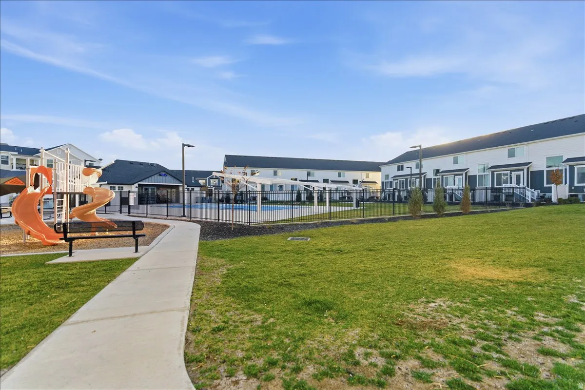 Expansive green space featuring a concrete pathway, a playground with slides and climbing structures, and a swimming pool with a surrounding fence