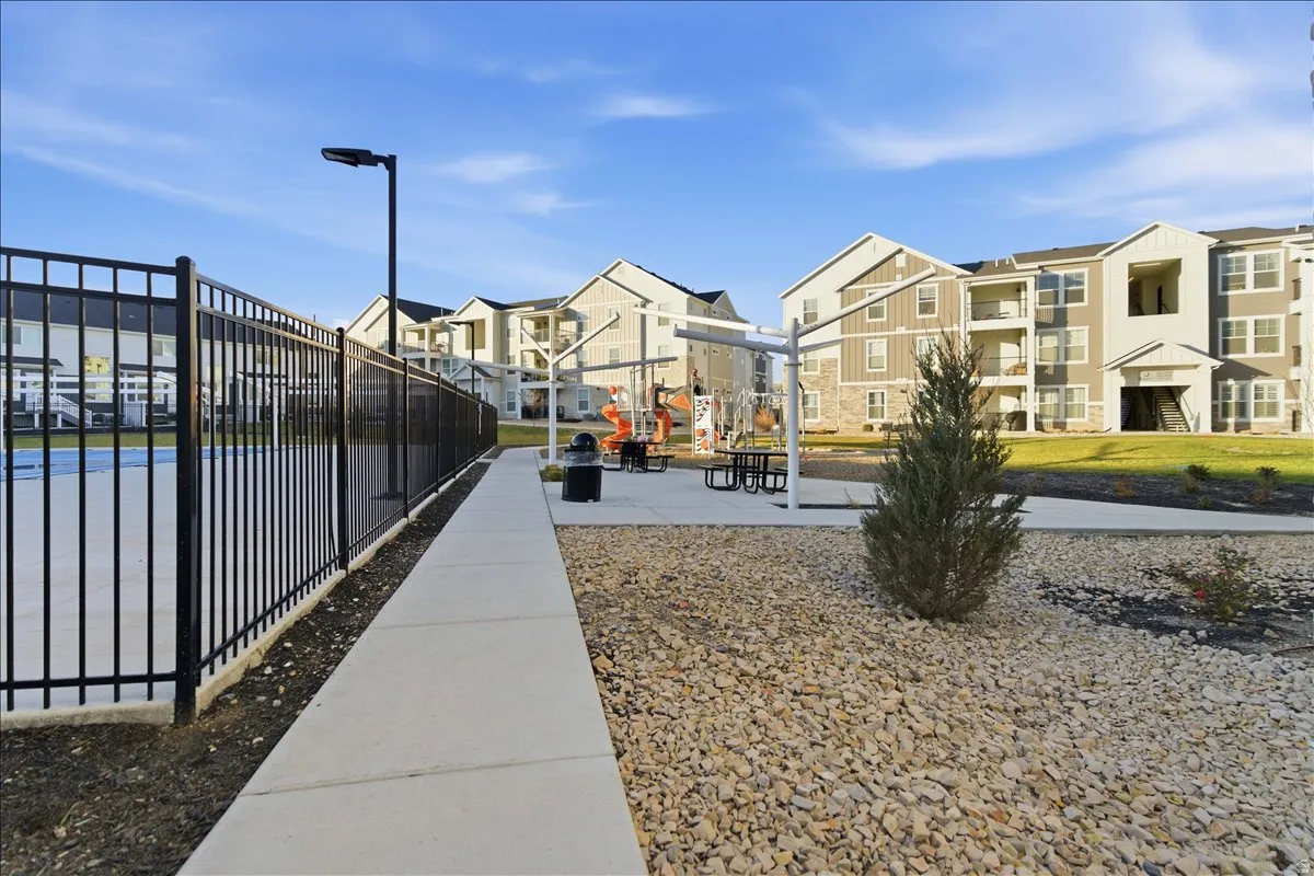 Outdoor recreational area featuring a black metal fence, concrete pathway, playground equipment, and picnic tables under a pergola