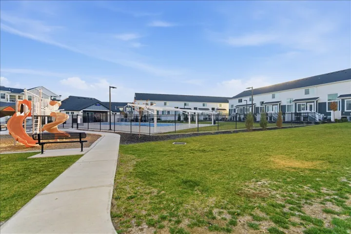 Expansive green space featuring a concrete pathway, a playground with slides and climbing structures, and a swimming pool with a surrounding fence