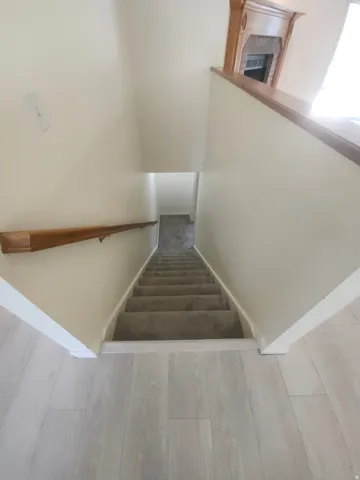 CStairs to basemeent. Carpeted staircase with a wood handrail, light wood-finish flooring, and a decorative fireplace mantel