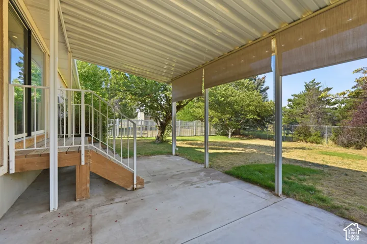 Covered concrete patio featuring a metal roof, white metal railing, and wood-finish steps, overlooking a lawn enclosed by metal fencing