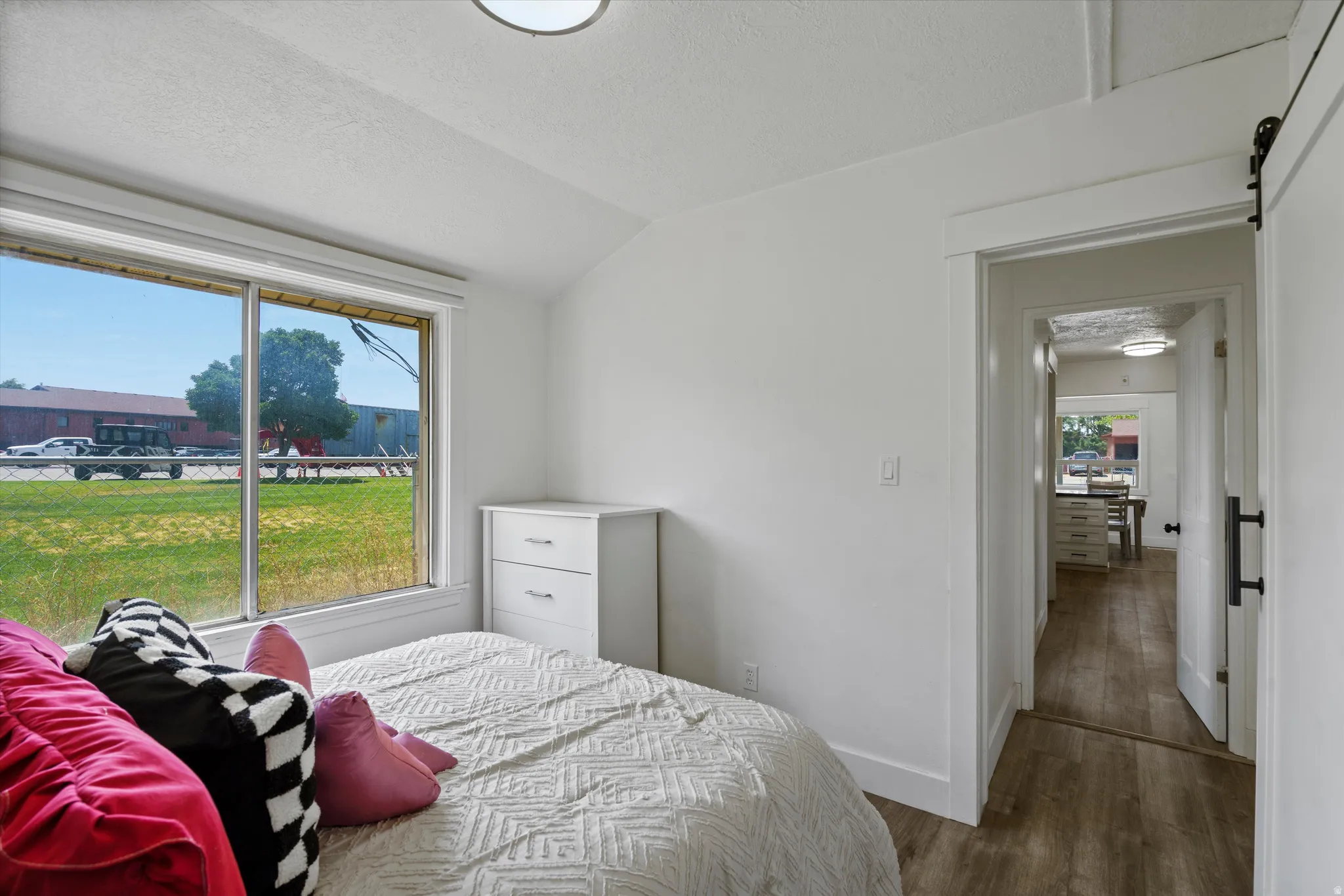 Bright bedroom featuring a large window, vaulted ceiling, wood-finish flooring, and a contemporary sliding barn door
