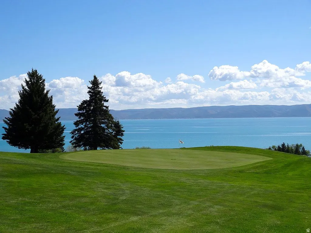 Expansive green golf course with a putting green and flagstick, featuring a large body of water and distant mountains under a blue sky with clouds