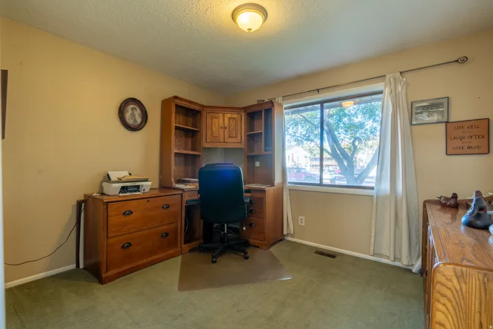 Carpeted room featuring a large window with white curtains, a built-in corner desk unit, and tan painted walls