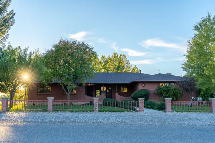 Brick exterior residence with a dark shingle roof, established landscaping, and a front yard enclosed by a decorative metal fence with brick pillars