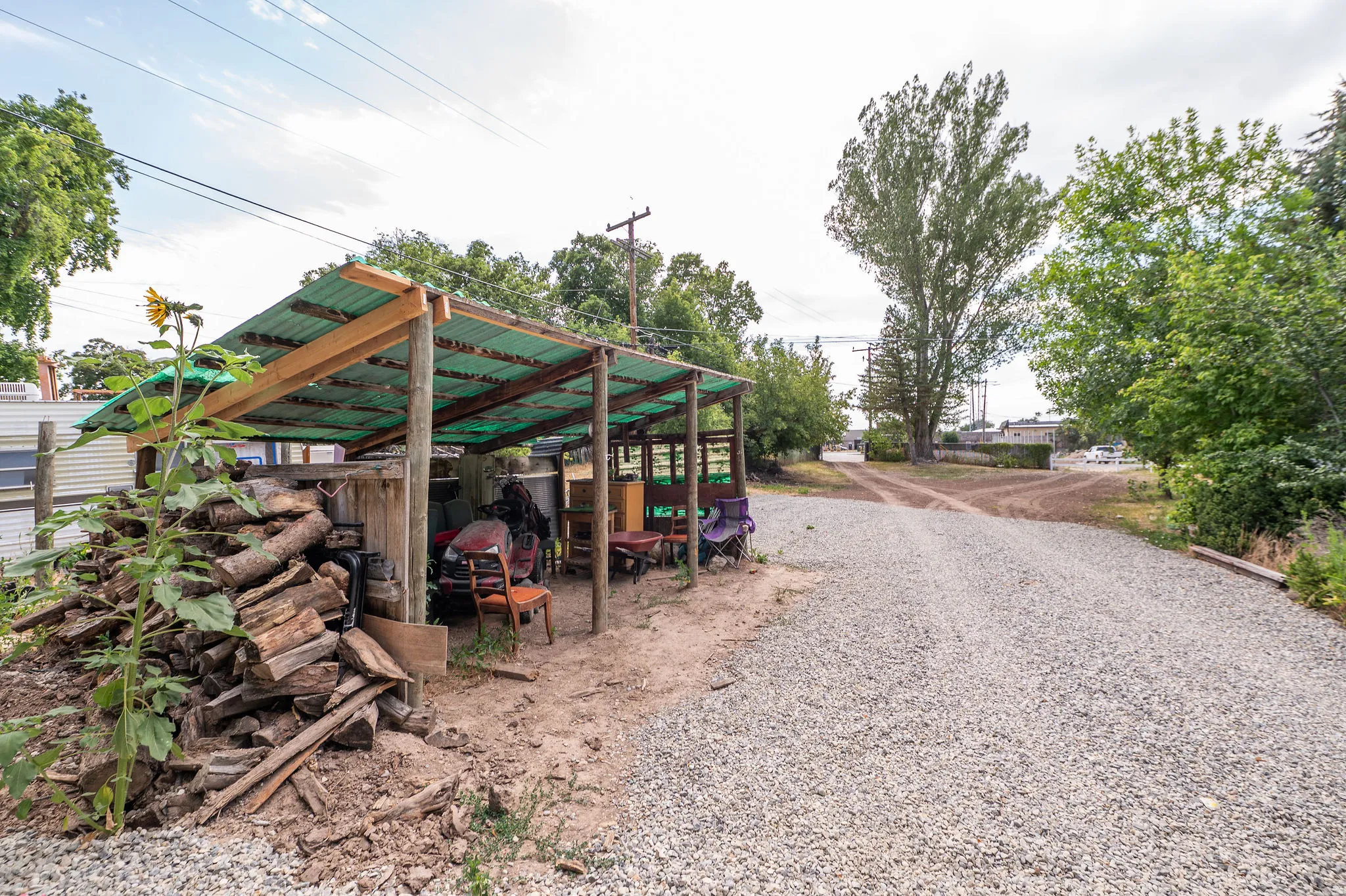Gravel driveway leading to a rustic open-sided shed with a corrugated green roof and wood framing, providing covered storage
