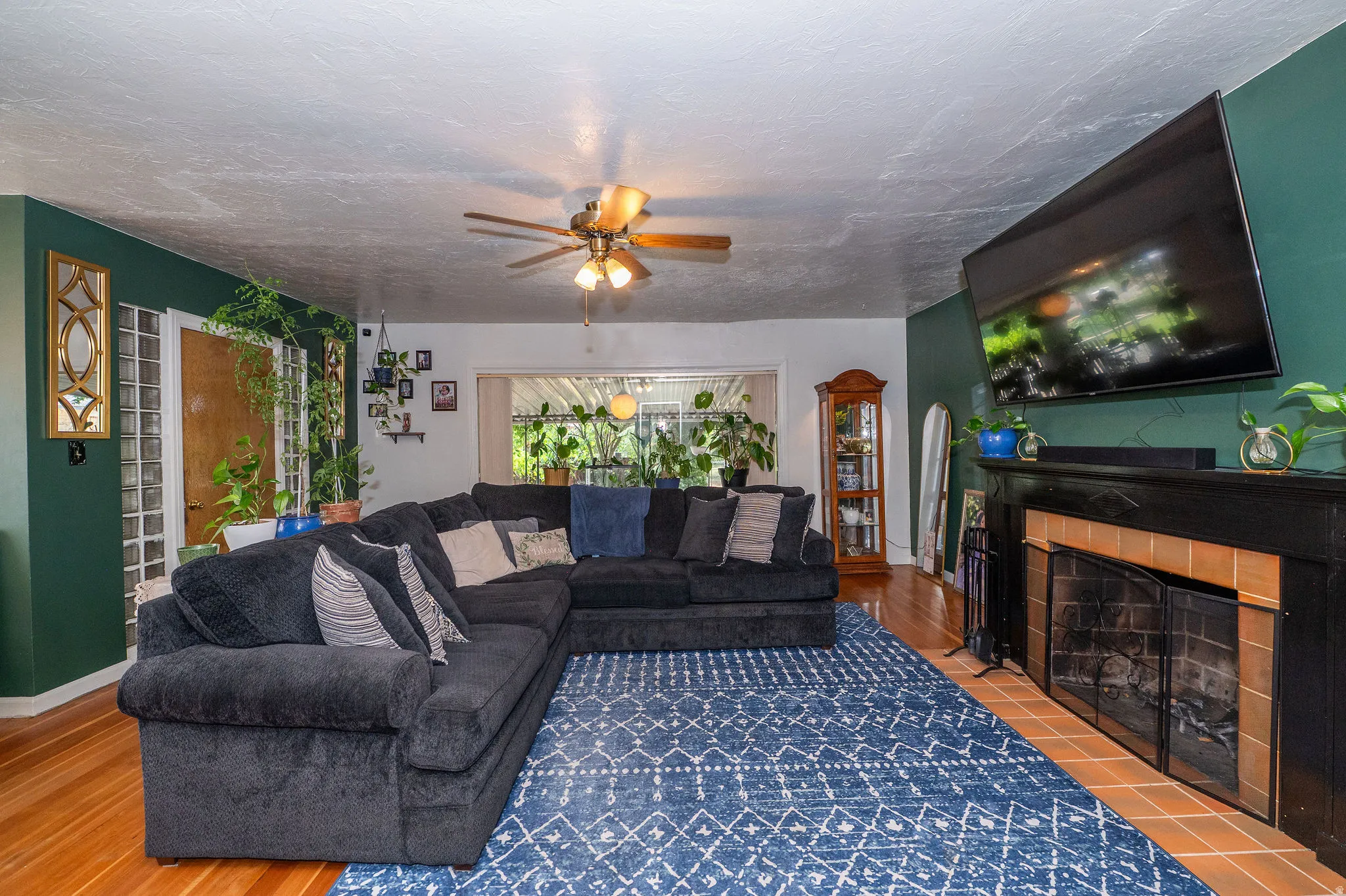 Living area featuring wood-finish flooring, a fireplace with a black mantel and terracotta tile surround, a ceiling fan, and a large window