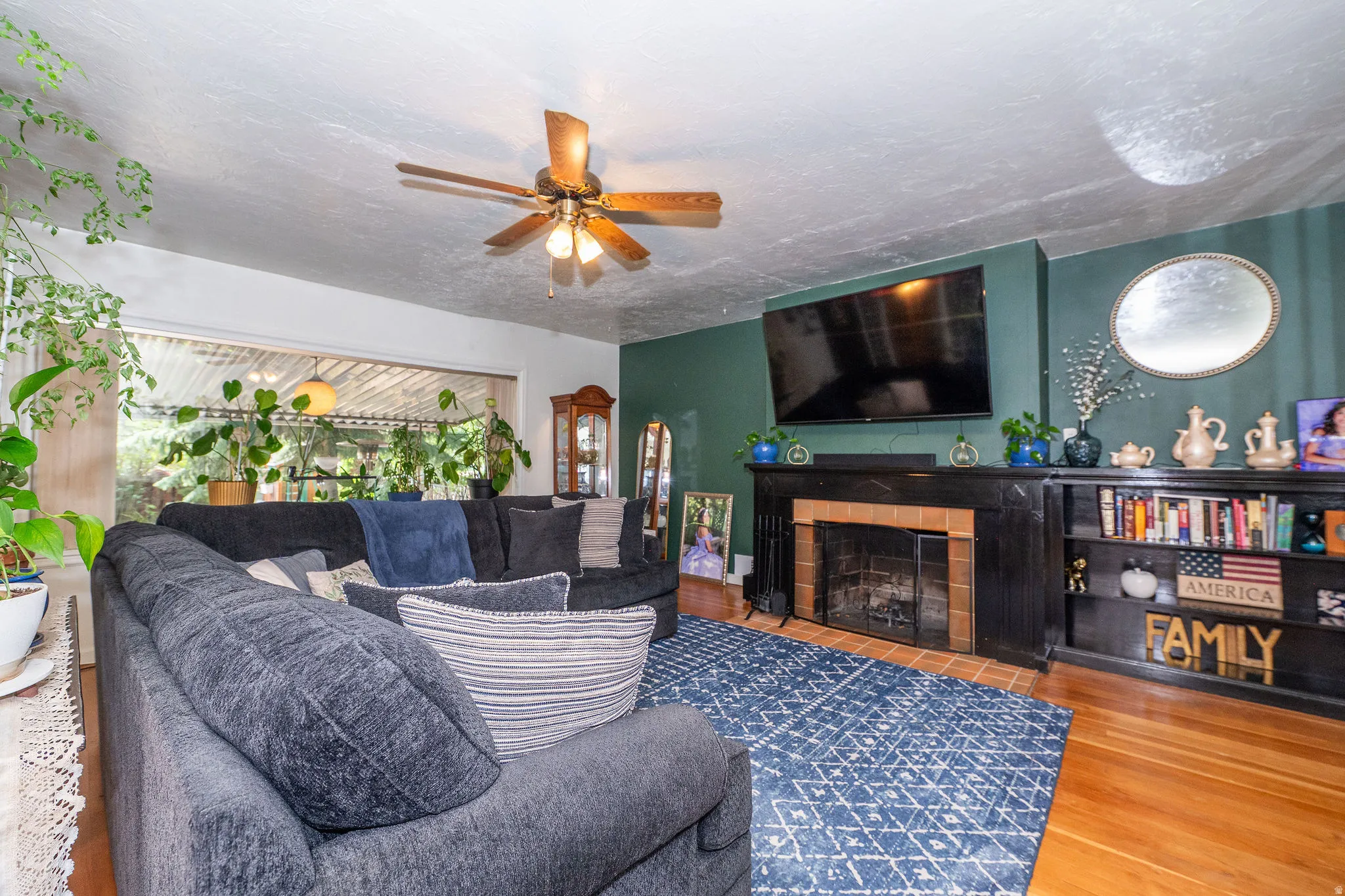 Living space featuring a fireplace with a brick surround and black mantel, built-in shelving, wood-finish flooring, and a ceiling fan