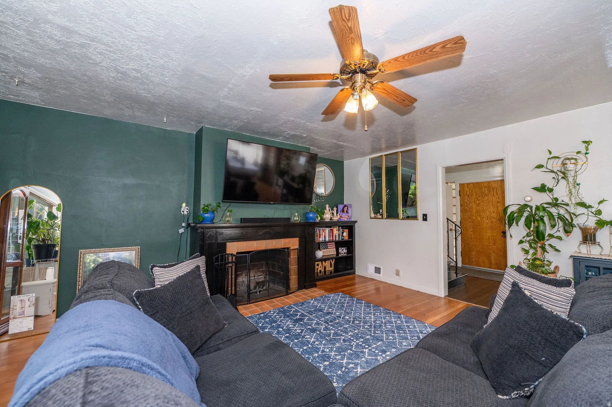 Living room featuring wood-finish flooring, a ceiling fan with integrated lighting, and a fireplace with a brick surround and black mantel