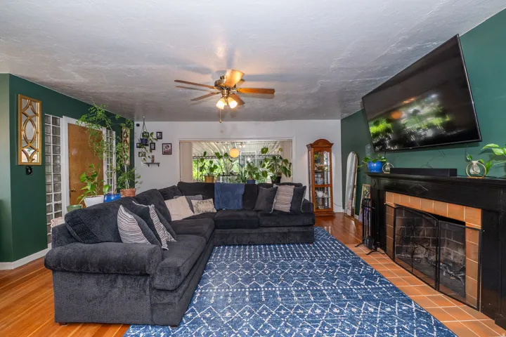 Living area featuring wood-finish flooring, a fireplace with a black mantel and terracotta tile surround, a ceiling fan, and a large window