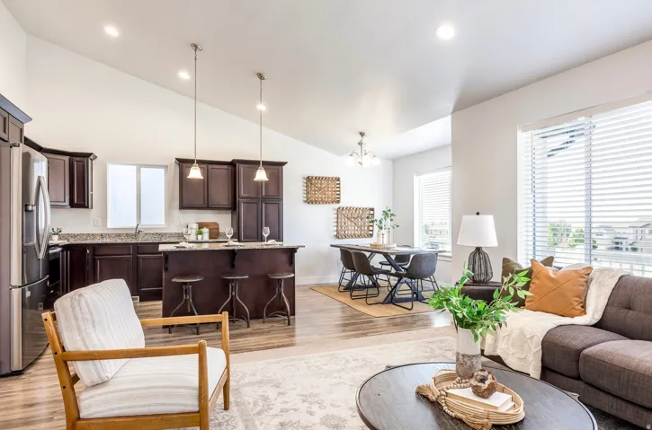 Open-concept living space featuring vaulted ceilings, wood-finish flooring, a kitchen island with pendant lighting, dark wood cabinetry, and large windows with blinds