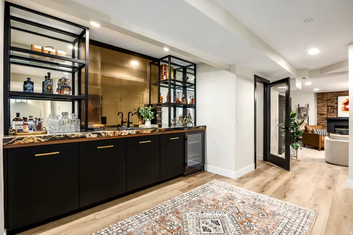 Built-in wet bar featuring black cabinetry with gold-tone hardware, a patterned countertop, a gold-toned mirrored backsplash, and open shelving