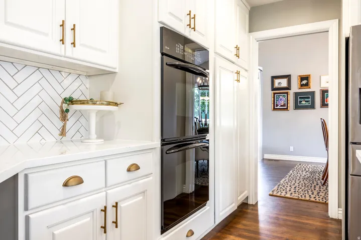 Built-in double oven with white shaker-style cabinetry, gold-finish hardware, white herringbone tile backsplash, and light-tone countertops
