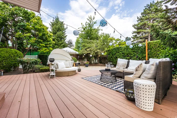 Expansive wood-finish deck with string lighting, surrounded by mature trees and manicured hedges, providing a private outdoor area