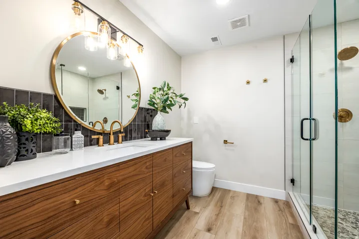 Bathroom featuring a wood-finish vanity with a white countertop, gold-finish fixtures, a circular mirror, and a glass-enclosed shower with tile surround