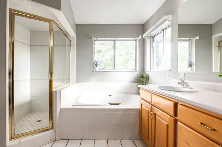 Bathroom featuring a built-in tub, separate shower with brass-finish enclosure, dual pane windows, a wood-finish vanity with an integrated sink, and white tile flooring