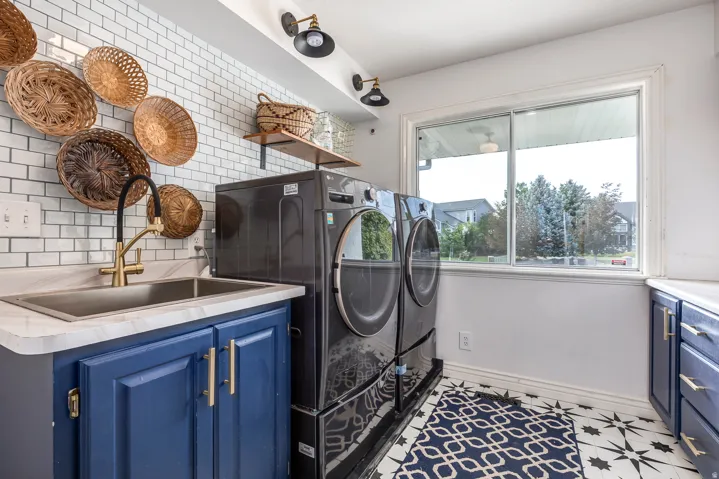 Laundry room featuring a single basin sink with a gold-tone faucet, blue cabinetry with gold-tone hardware, white subway tile backsplash, and patterned floor tile