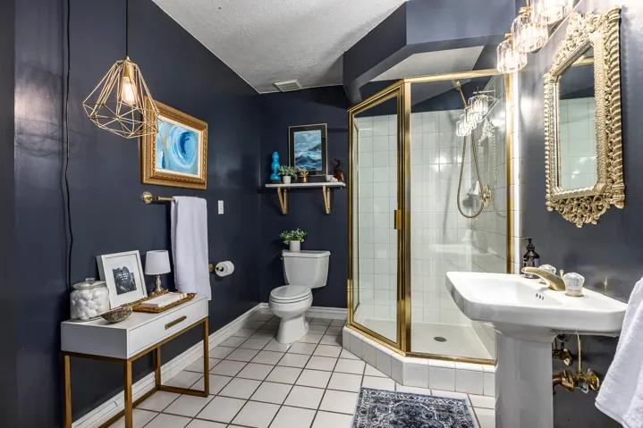 Bathroom featuring a corner shower with a brass-framed glass enclosure, white square tile flooring, a pedestal sink with brass-finish fixtures, and a decorative gold-framed mirror