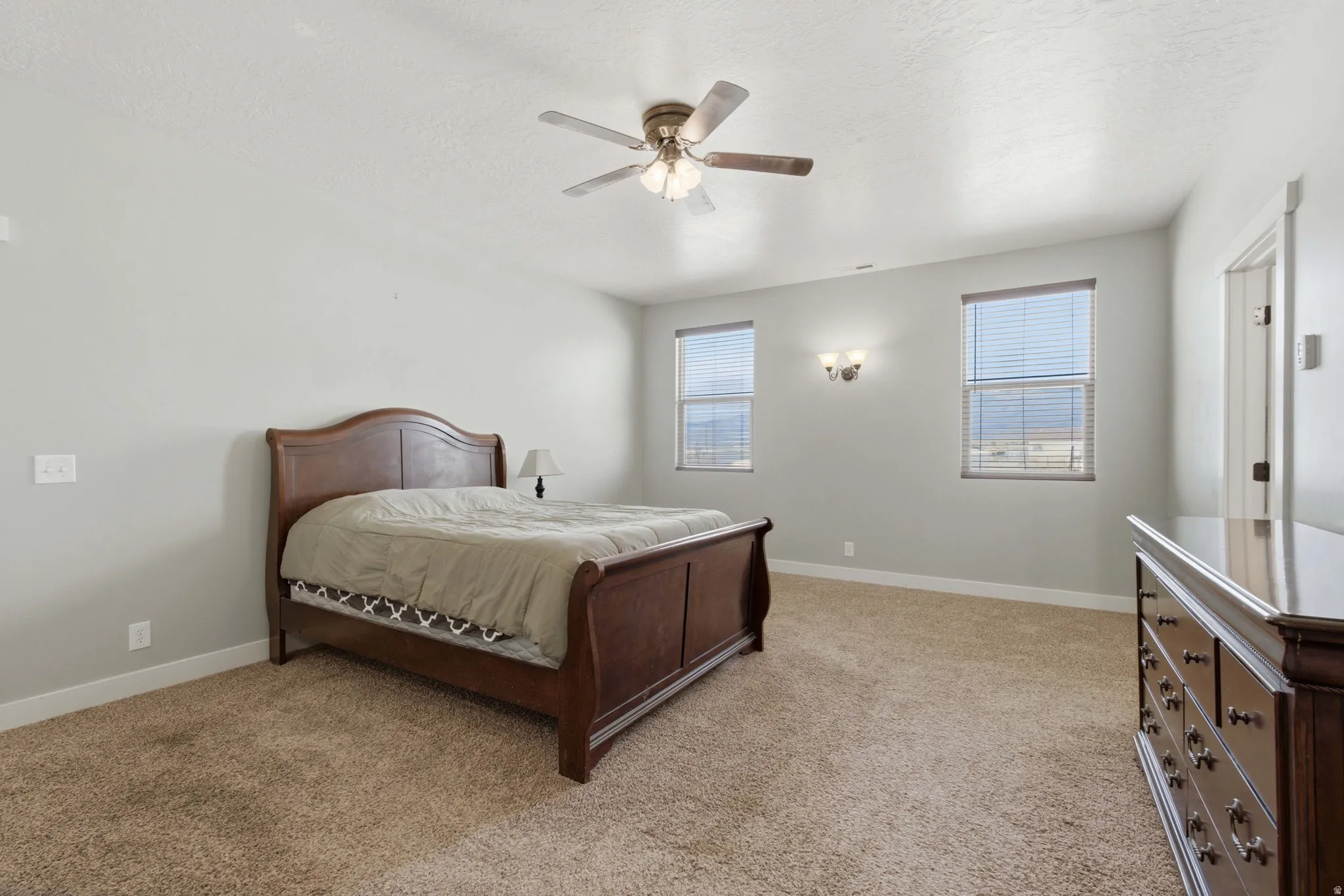 Carpeted room featuring light gray walls, two windows with horizontal blinds, a ceiling fan with integrated lighting, and a wall-mounted light fixture