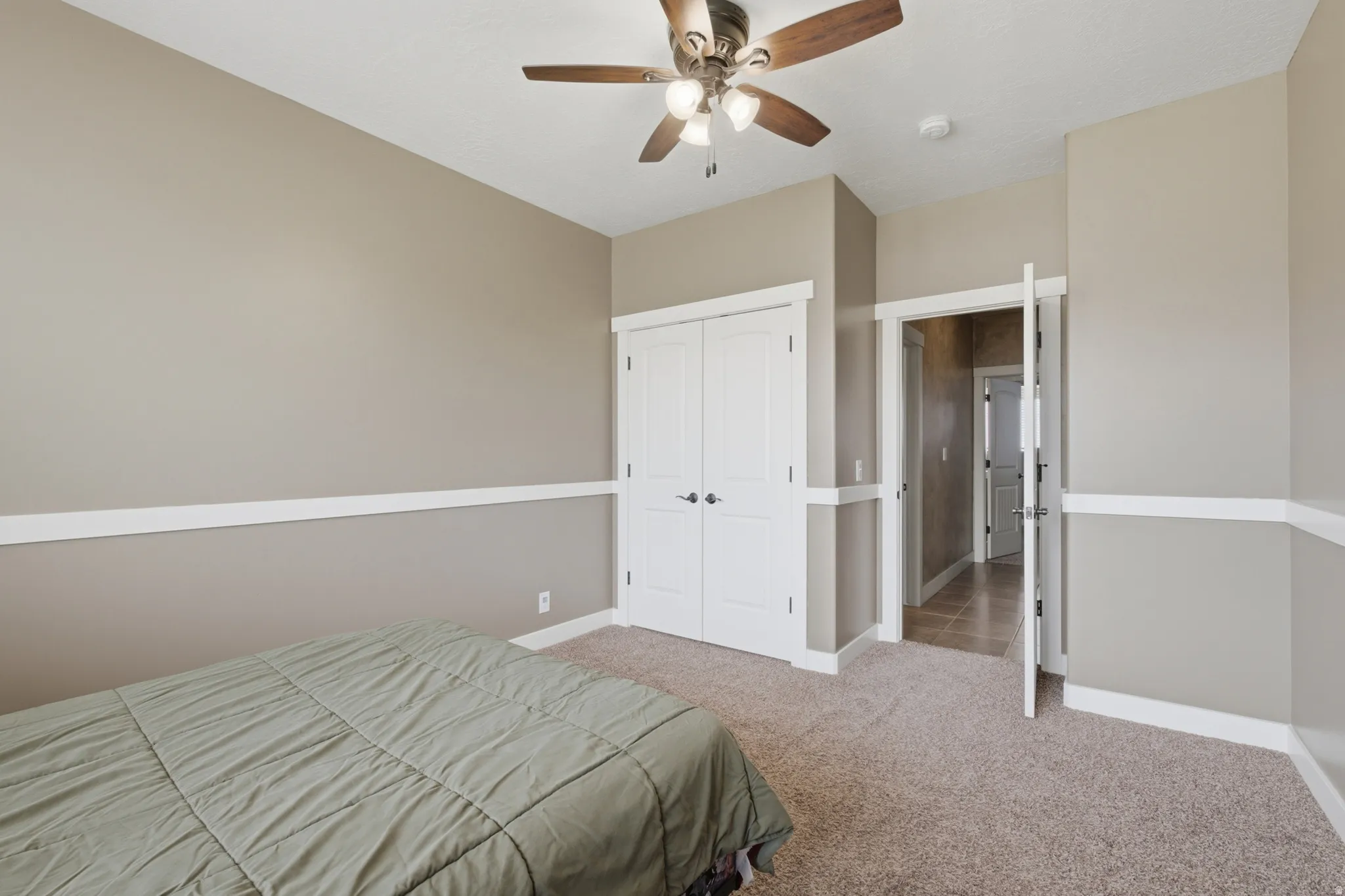 Bedroom featuring neutral wall paint with white chair rail molding, a ceiling fan with integrated lighting, carpeted flooring, and a pair of white bifold closet doors