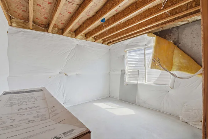 Unfinished basement space featuring exposed wood joists, insulation, a window with venetian blinds, concrete flooring, and wrapped walls