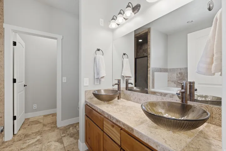 Dual vessel sinks set on a stone-look countertop with wood cabinetry, large vanity mirror, and tile flooring