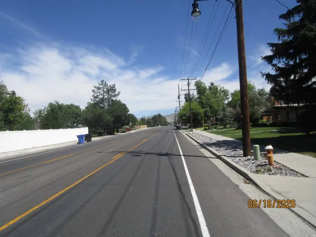 Paved roadway with painted lane markings bordered by a concrete sidewalk, mature trees, and a white privacy fence
