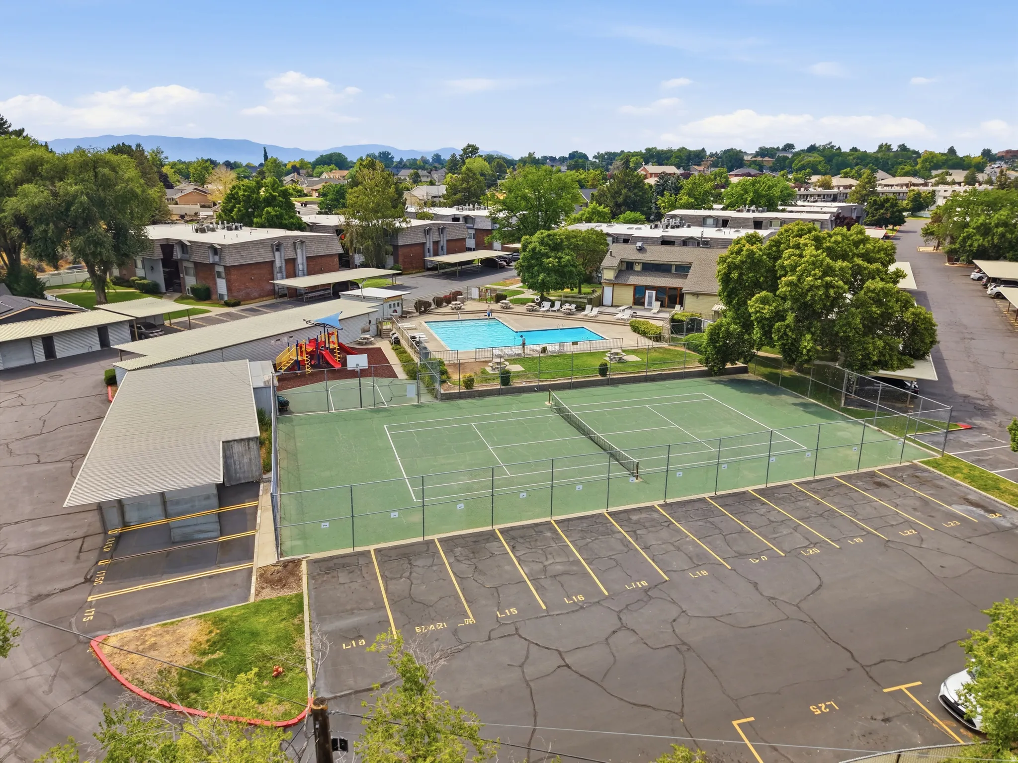 Outdoor tennis court with chain-link fencing, adjacent to a swimming pool, covered parking structures, residential buildings, and a visible playground area