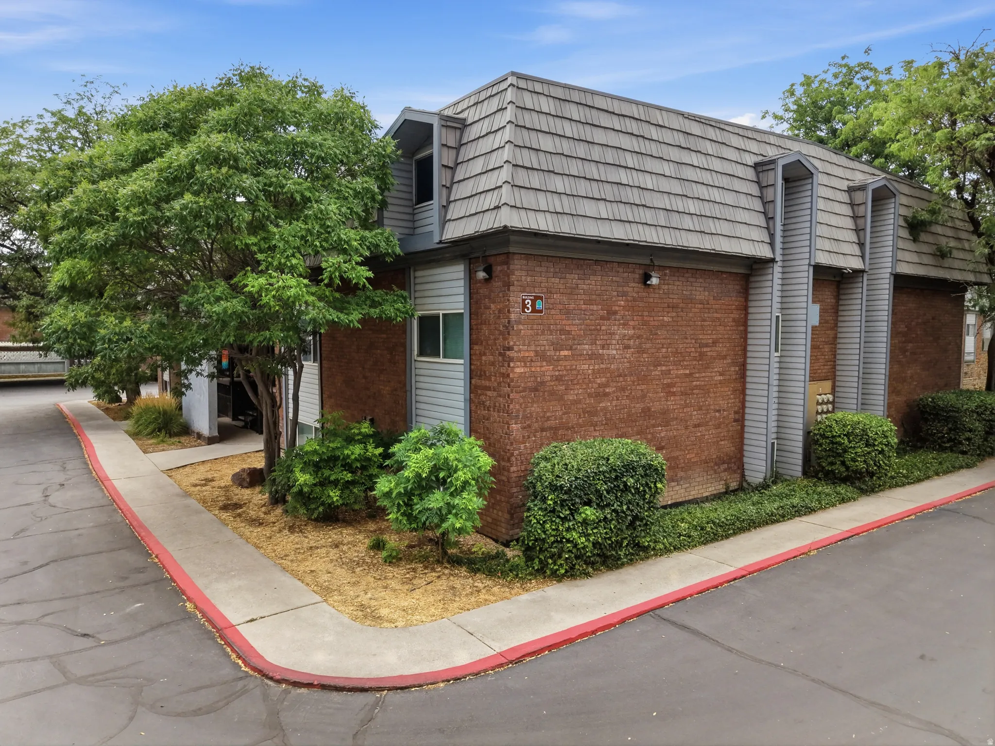 Multi-unit building featuring brick and siding exterior, a textured roof, mature trees, and established landscaping