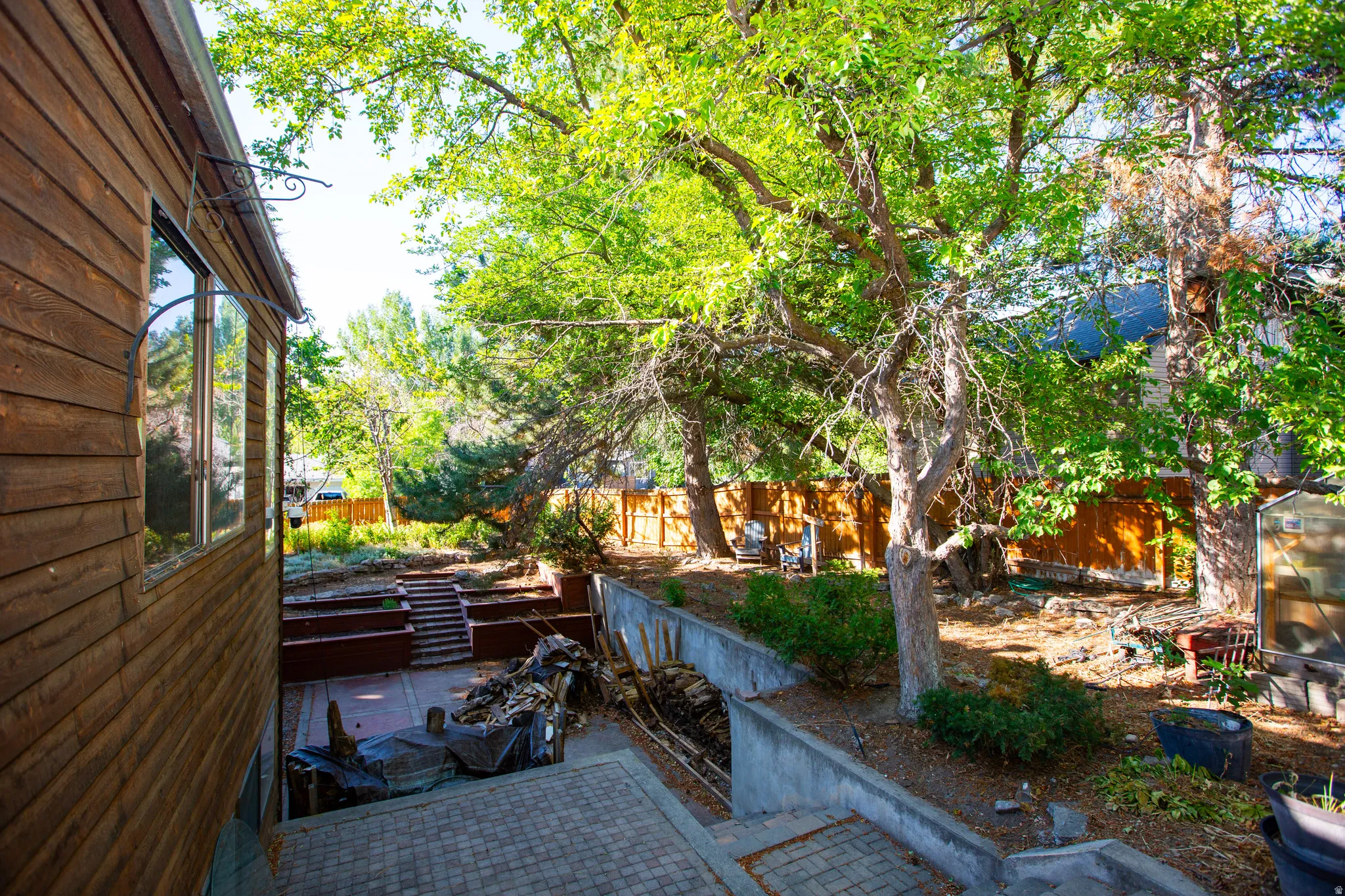 Wood-paneled exterior featuring a large window, paver patio, tiered retaining walls, and mature trees