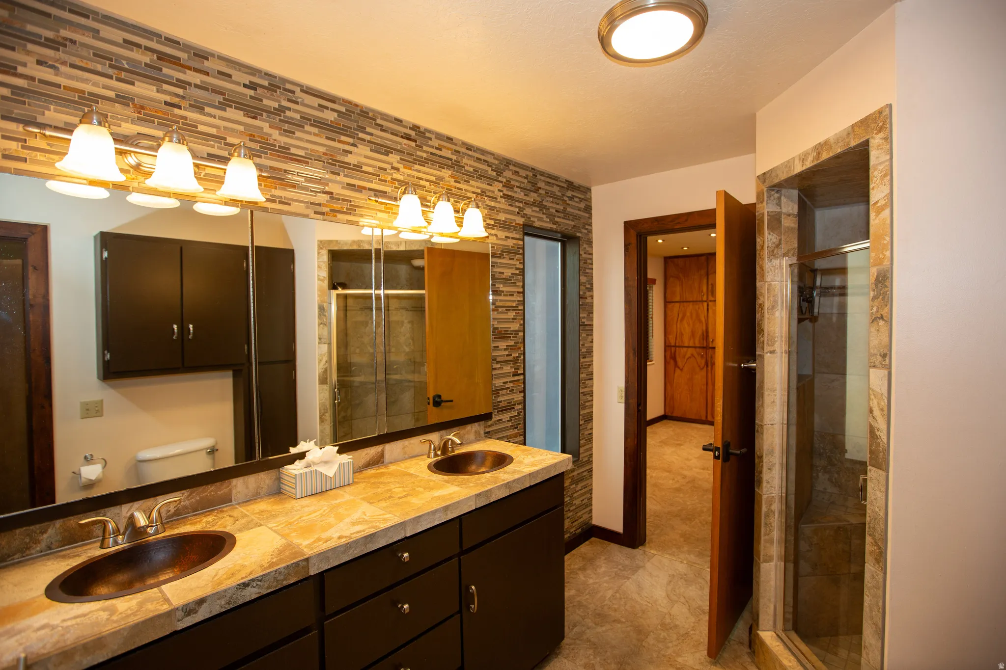 Master bathroom featuring a dual vanity with a tile countertop and copper-finish vessel sinks, a full-width mirror, a tiled accent wall, and a glass-enclosed shower with tile surround