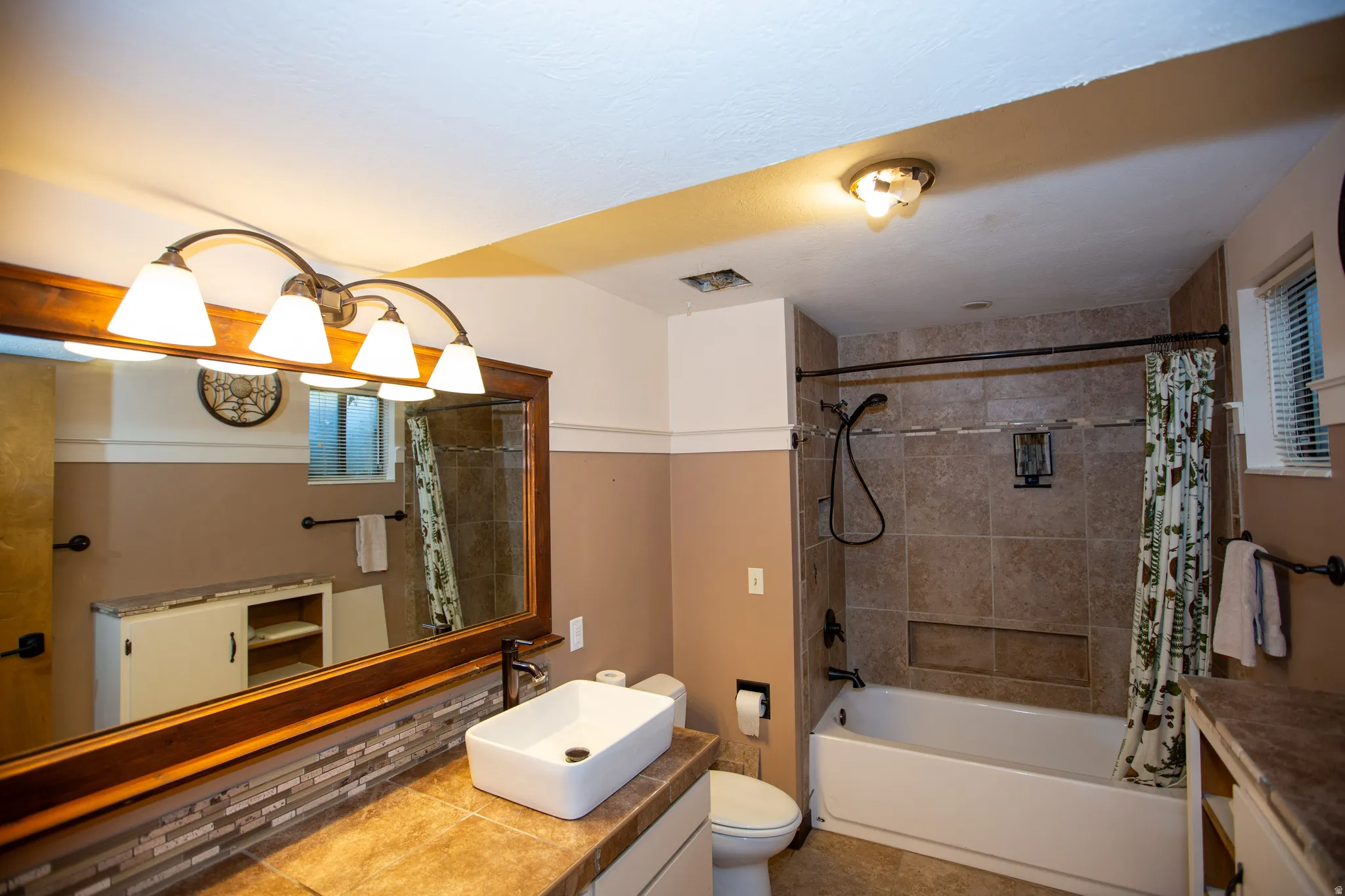 Full bathroom featuring a built-in tub with tile surround, a vessel sink on a tiled countertop, a wooden framed mirror, and a four-light vanity fixture