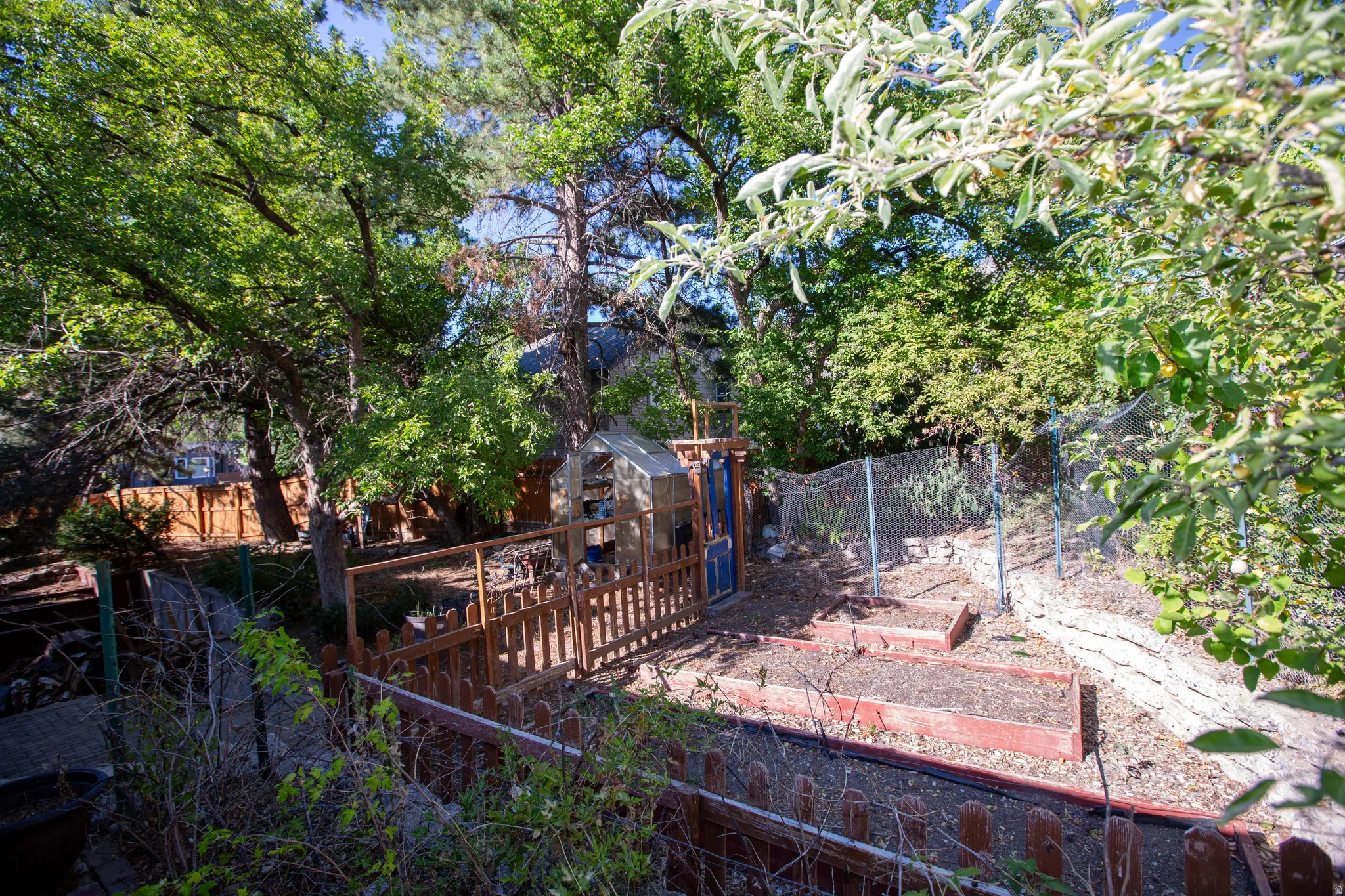 Lush, treed backyard featuring a fenced garden area, raised garden beds, wood fencing, and a small utility structure