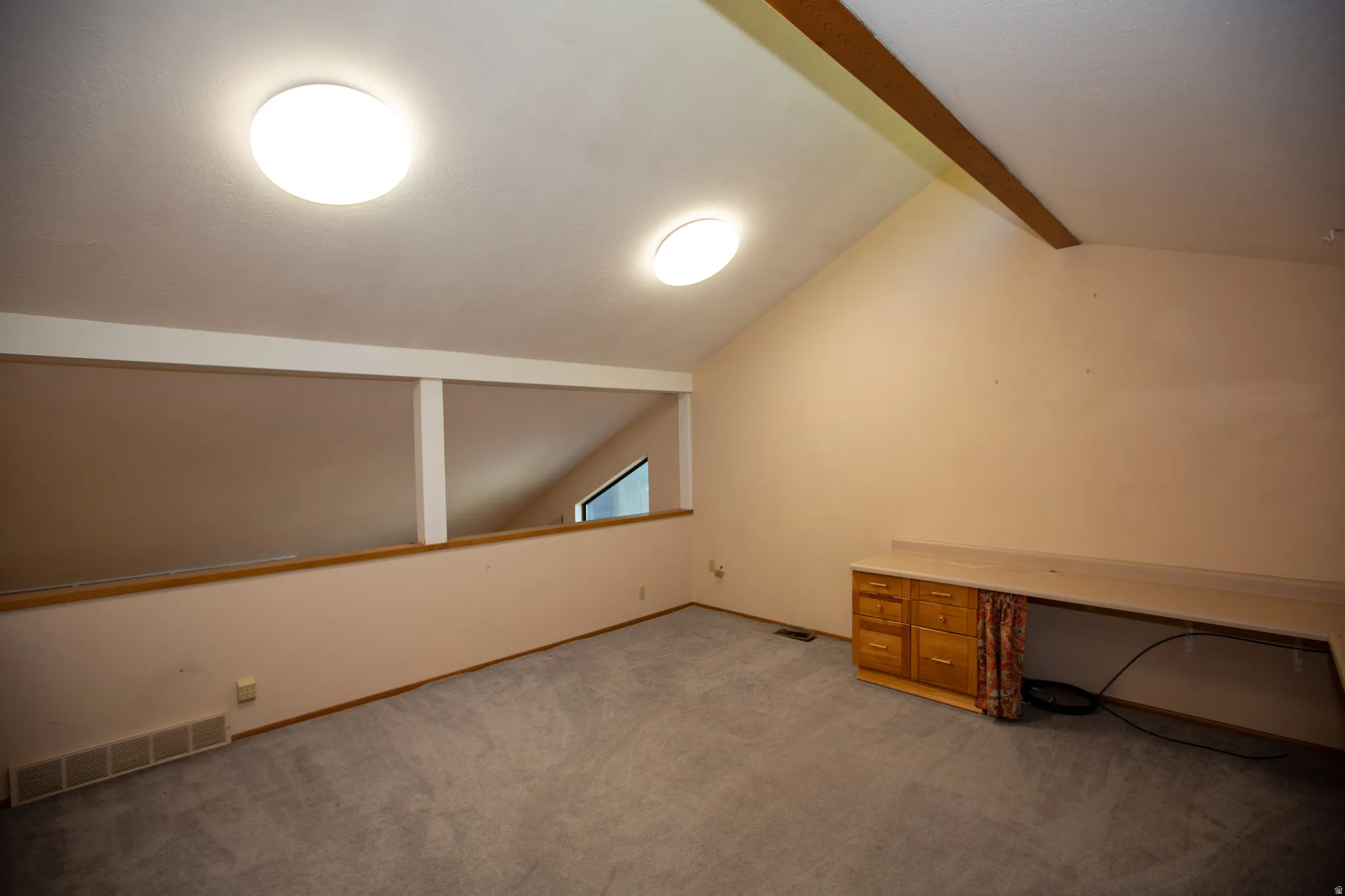 Carpeted room featuring a built-in wood desk with drawers, an angled ceiling, and a large window providing natural light