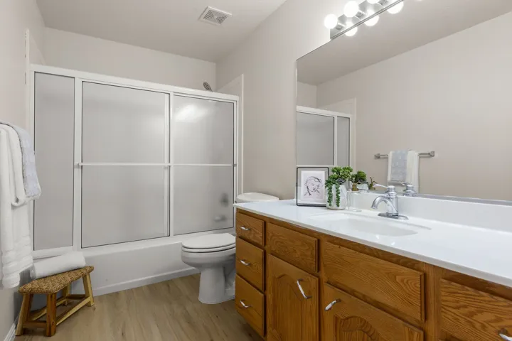 Wood-finish flooring, full-width vanity mirror, chrome fixtures, built-in wooden cabinetry, and a shower-tub combination with frosted sliding doors