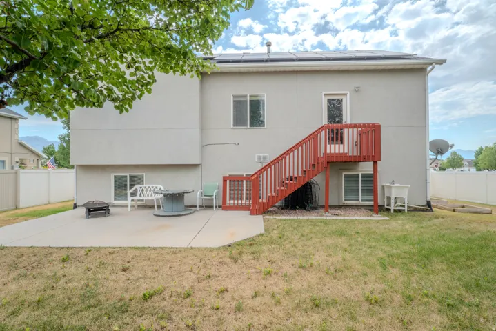 Rear exterior featuring a large concrete patio, red-stained wooden deck and stairs, multiple windows, and solar panels