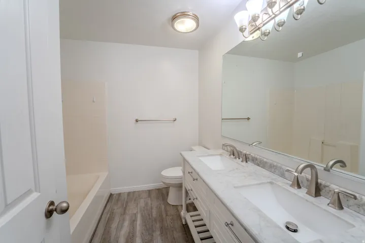 Bathroom featuring a dual vanity with a stone countertop, brushed nickel fixtures, a wide mirror, wood-finish flooring, and a tub-shower combination with subway tile surround