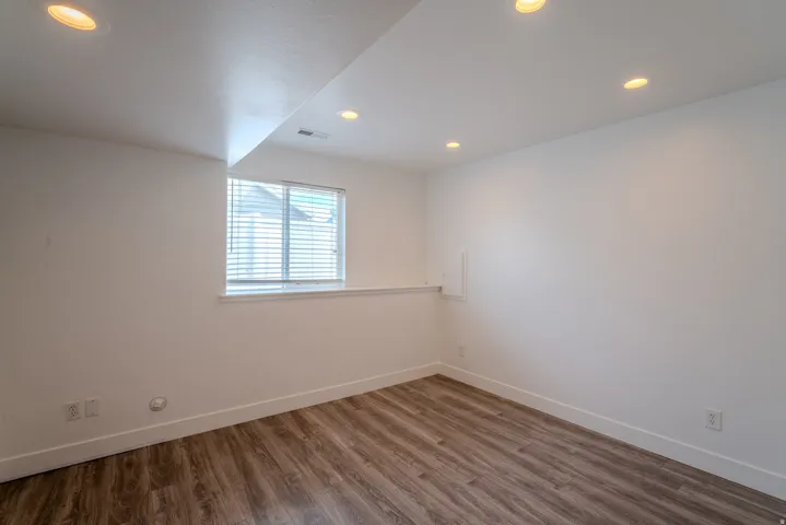 Spacious room featuring neutral wall tones, recessed lighting, wood-finish flooring, a window with horizontal blinds, and white baseboards