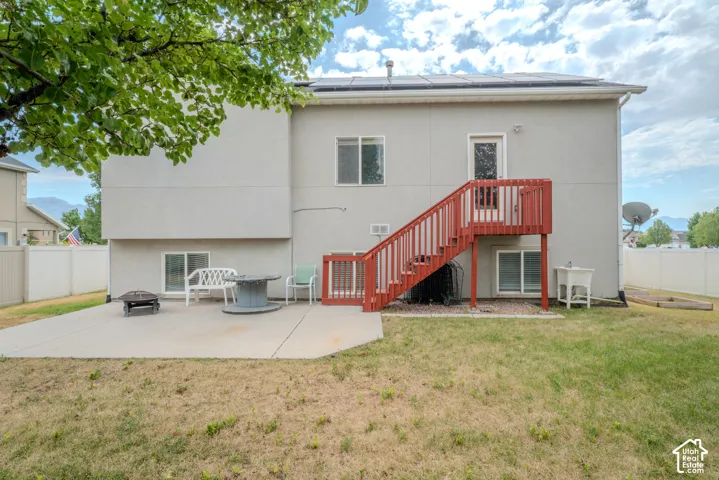 Rear exterior featuring a large concrete patio, red-stained wooden deck and stairs, multiple windows, and solar panels