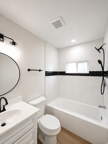 Bathroom featuring a white vanity with integrated sink, matte black fixtures, wood-finish flooring, and a tub-shower combination with textured white tile and a black accent band