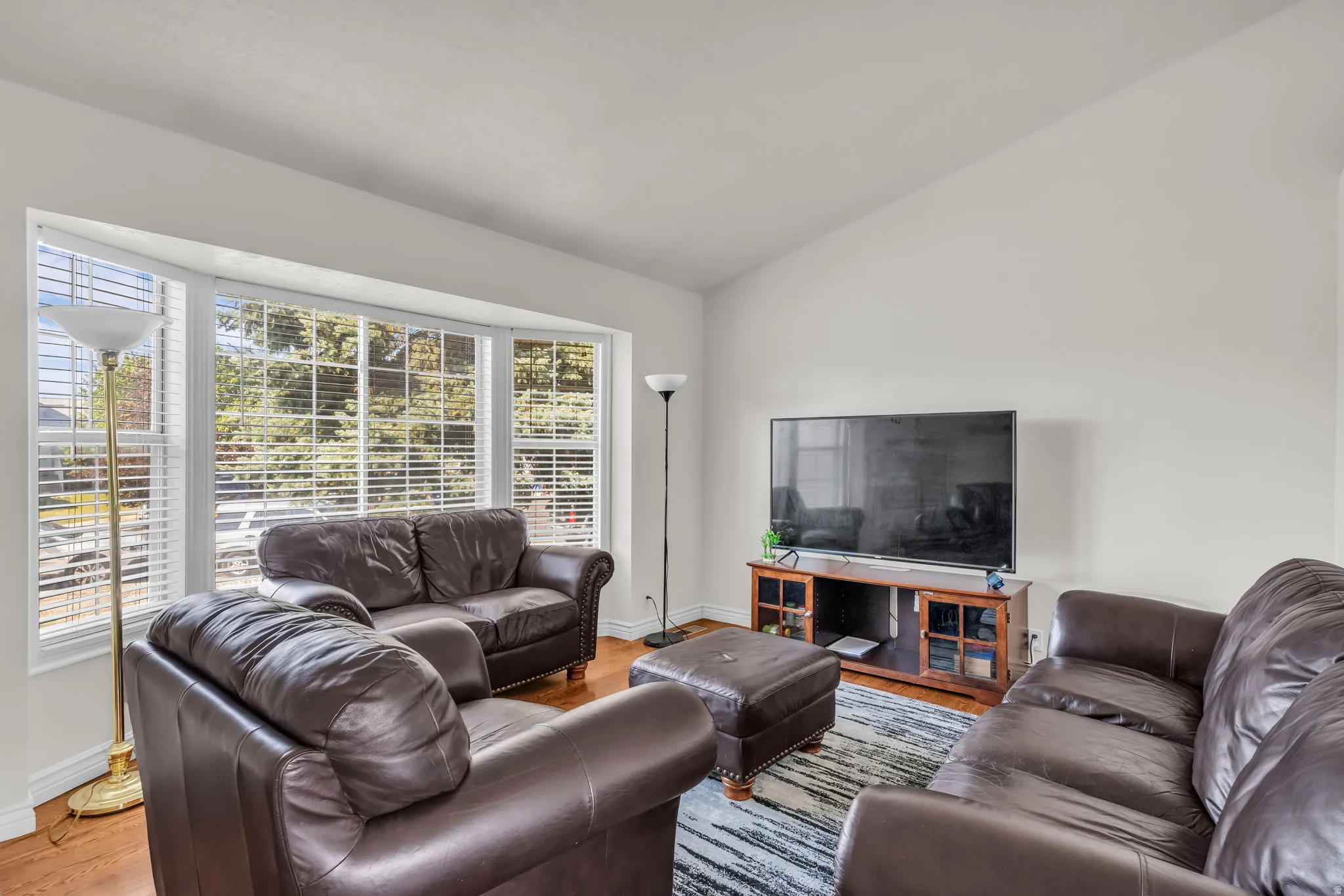Living area featuring a prominent bay window with white blinds, vaulted ceiling, and wood-finish flooring