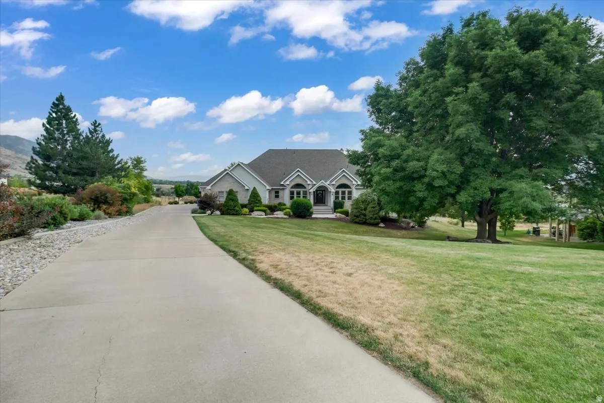 Paved driveway leading to a residence featuring a traditional facade, mature trees, manicured landscaping, and a mountain backdrop