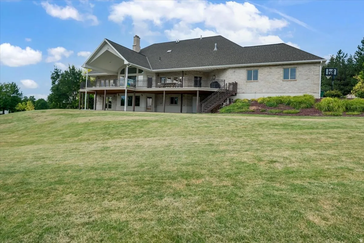Expansive rear elevation featuring a light-colored brick exterior, a covered upper-level balcony with metal railings, a lower-level patio, and a tiered wooden deck with stairs descending to the lawn