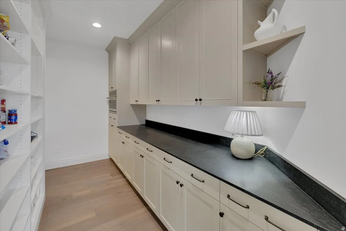 Expansive pantry featuring custom white cabinetry with dark hardware, dark stone countertops, wood-finish flooring, and open shelving