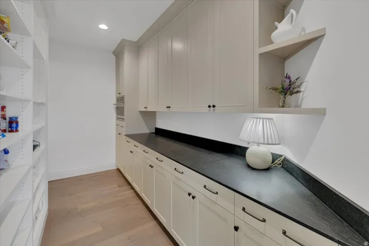 Expansive pantry featuring custom white cabinetry with dark hardware, dark stone countertops, wood-finish flooring, and open shelving