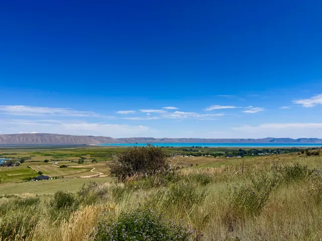 Expansive natural landscape featuring a prominent blue body of water, distant mountain ranges, and verdant fields