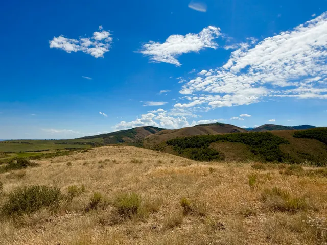 Expansive rural landscape featuring rolling hills, natural vegetation, and a clear sky with scattered clouds