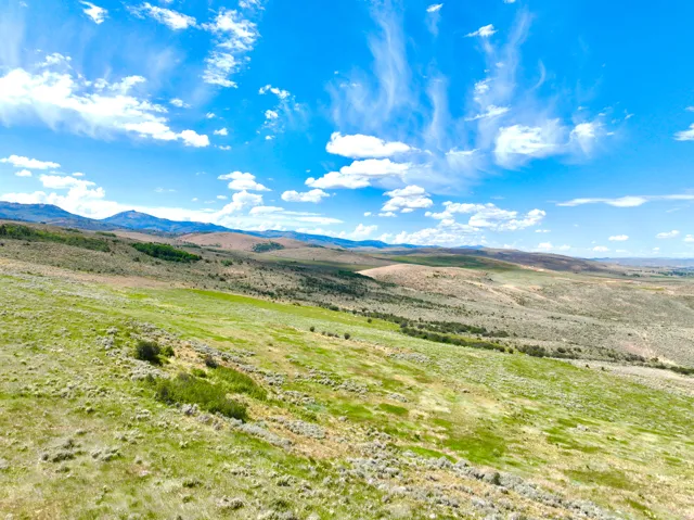 Expansive rolling landscape featuring varied green and brown vegetation, distant mountains, and a vibrant blue sky with scattered clouds
