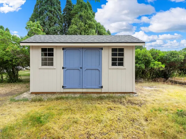 Detached storage shed featuring vertical panel siding, a shingle roof, blue double doors with black hardware, and two paneled windows